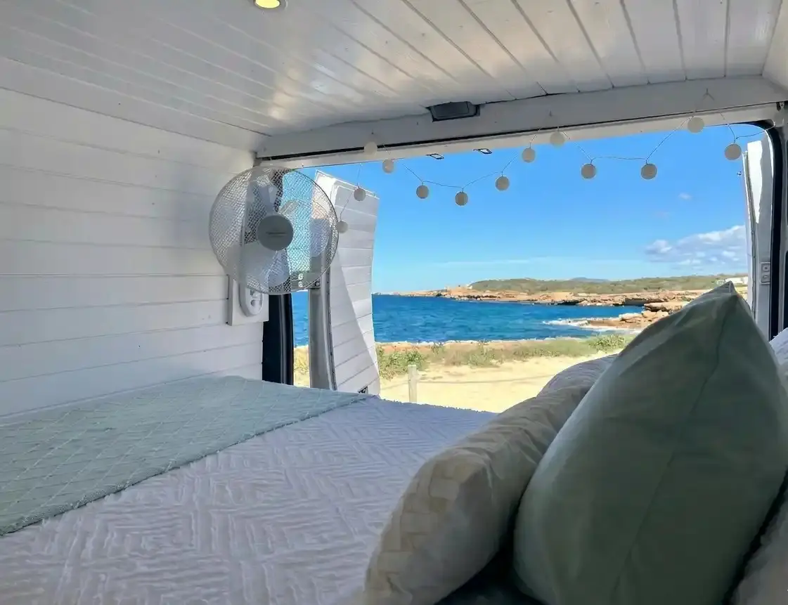 Cama de matrimonio en el interior de una furgoneta camper de alquiler en Ibiza, con las puertas traseras abiertas hacia una playa de aguas turquesas.