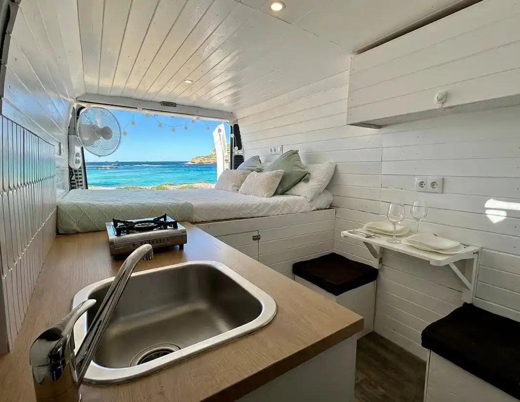 Interior de furgoneta camper de alquiler Peugeot Boxer en Ibiza, con cocina de madera, cama hecha y vistas al mar turquesa desde las puertas traseras abiertas.