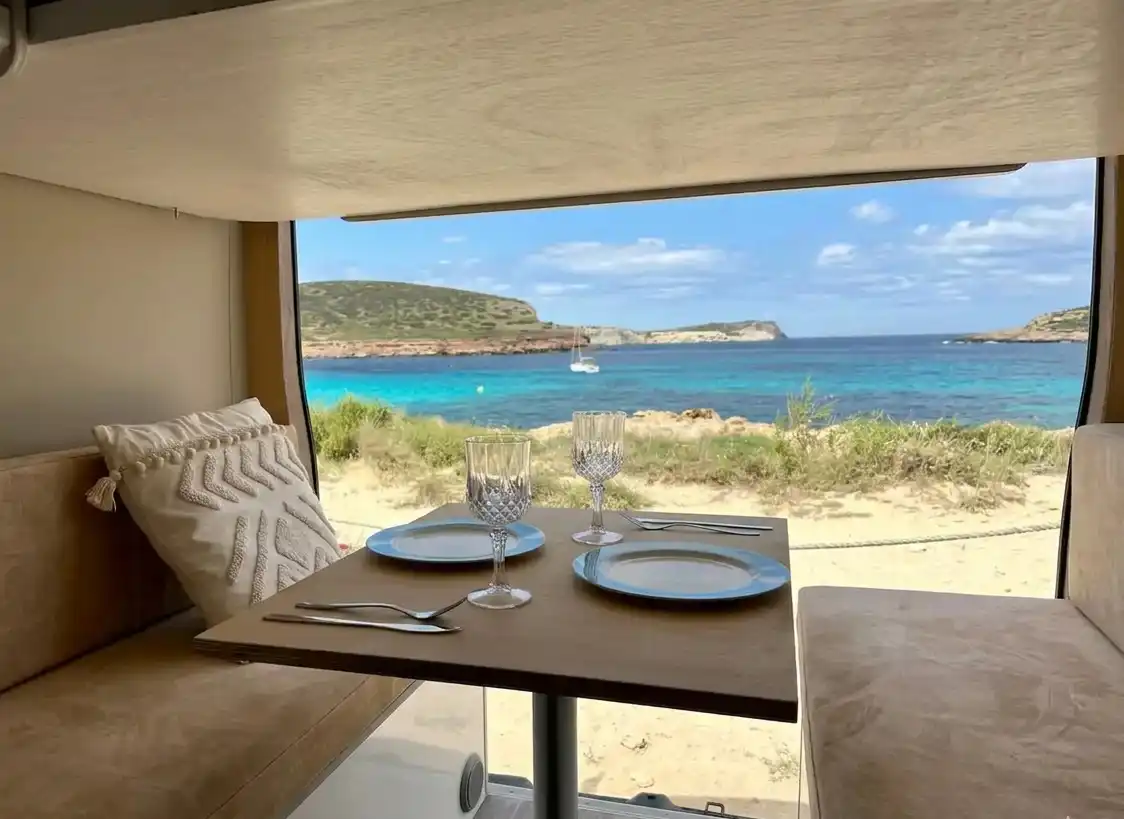 Salón comedor interior de camper Hyundai H350 con vistas a la playa de Cala Comte en Ibiza.